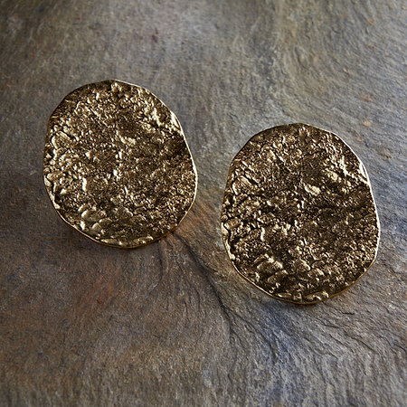 The Oya Hammered Earrings - Gold by nkuku, featuring textured irregular ovals and an artisanal finish, rest on a rough stone surface with green leaves partially visible on the left.