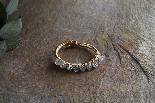 The Vani Labradorite Ring by nkuku, featuring labradorite stones and gold detailing, is coiled in a circle on a textured brown stone surface, with green leaves partially visible in the upper left corner.