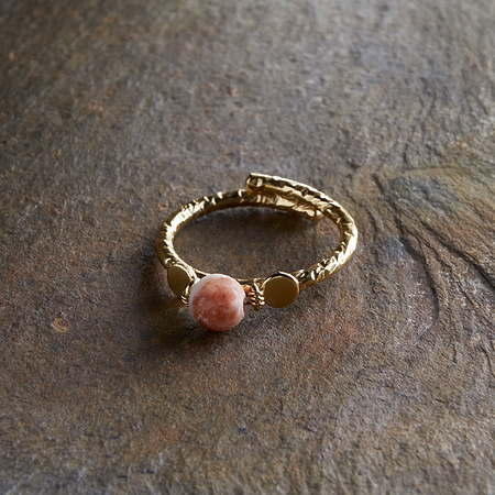 The Cerita Rhodochrosite Ring by nkuku, featuring a pink rhodochrosite stone and textured gold-plated band, is displayed on a rough brown stone with green leaves in the upper left corner.