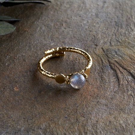 The Marai Labradorite Ring by nkuku features a textured gold-plated band with round details and a translucent labradorite centerpiece, displayed on a rough stone surface with green leaves in the upper left corner.