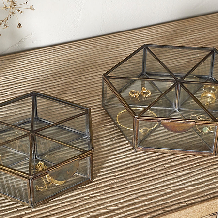 Two nkuku Sitara Jewellery Boxes in antique brass sit on a ribbed wooden surface. The smaller holds a gold bracelet and ring, the larger has earrings, rings, and a necklace. A glass vase with dried flowers is nearby.