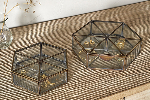 Two nkuku Sitara Jewellery Boxes in antique brass sit on a ribbed wooden surface. The smaller holds a gold bracelet and ring, the larger has earrings, rings, and a necklace. A glass vase with dried flowers is nearby.