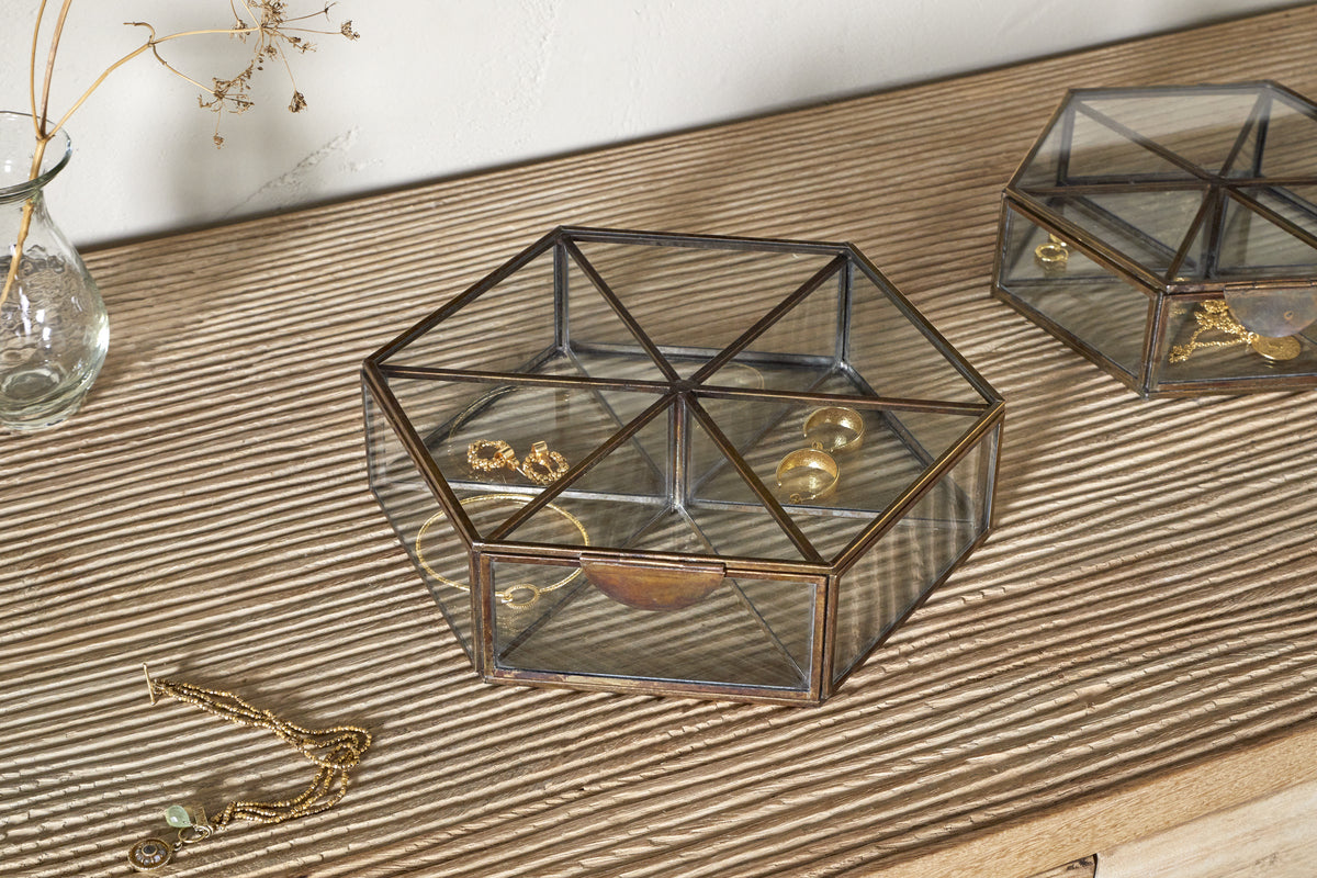 The Sitara Jewellery Box - Antique Brass by nkuku rests on a ribbed wooden surface, holding gold jewelry and a dish. Nearby are another similar box, scattered jewelry, and a small vase with dried flowers in the corner.