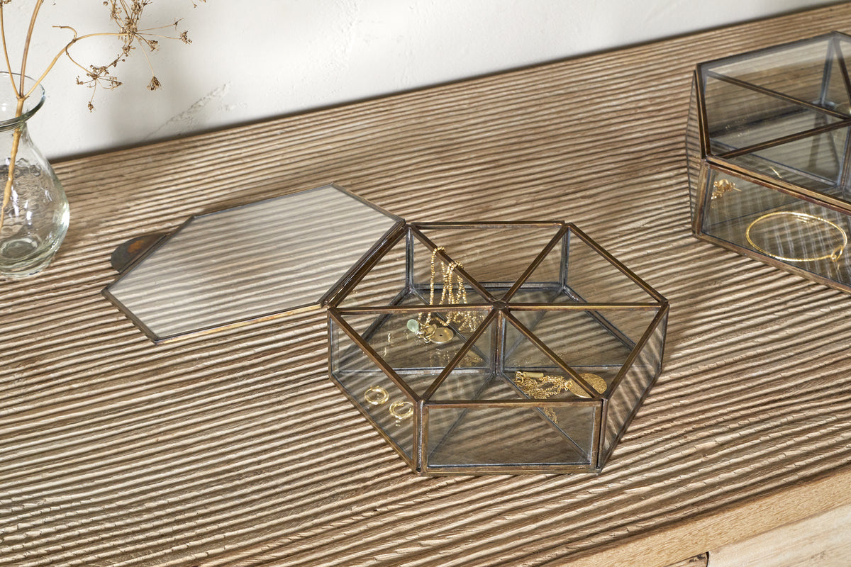 The Sitara Jewellery Box - Antique Brass by nkuku sits open on a ribbed wooden surface, displaying assorted gold rings and bracelets beside a small vase of dried flowers and another closed glass box.