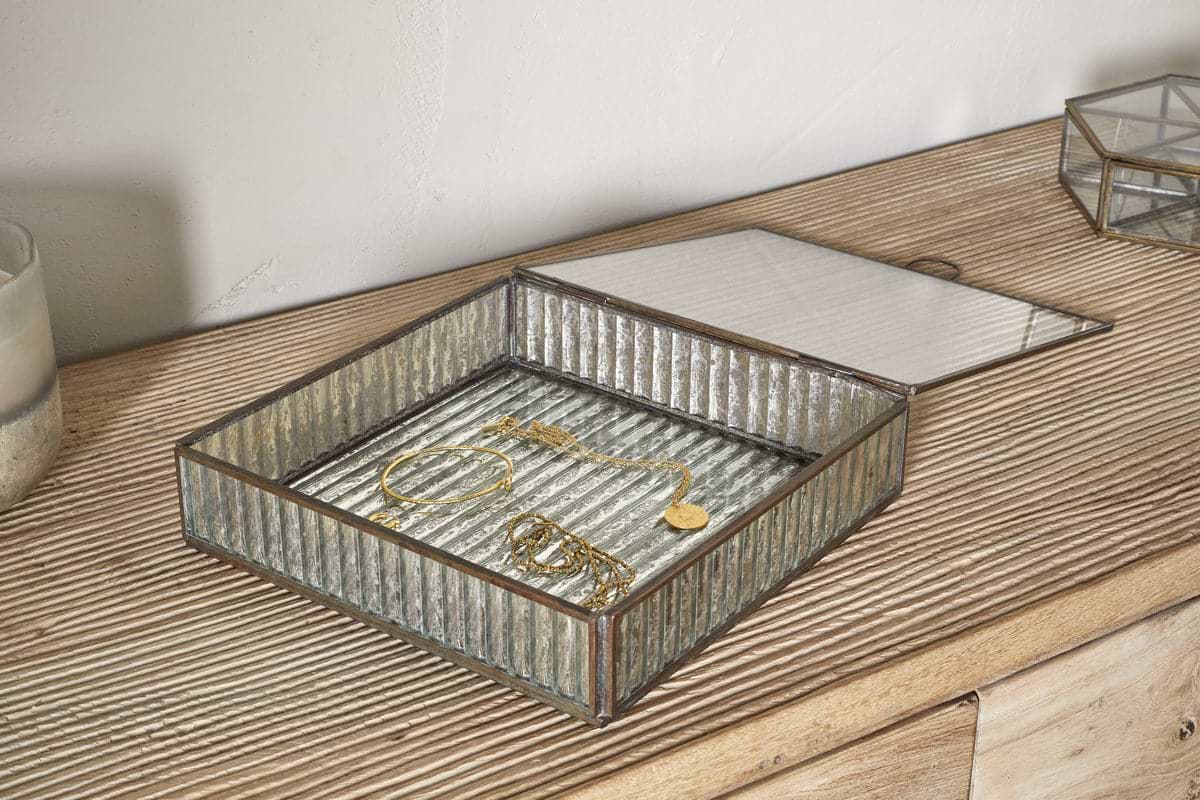 The Malita Jewellery Box - Antique Brass by nkuku sits open on a textured wooden dresser, displaying a gold bracelet, necklace, and earrings. Its ribbed glass lid is partially lifted, with a candle placed nearby.