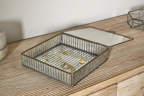 The Malita Jewellery Box - Antique Brass by nkuku sits open on a textured wooden dresser, displaying a gold bracelet, necklace, and earrings. Its ribbed glass lid is partially lifted, with a candle placed nearby.