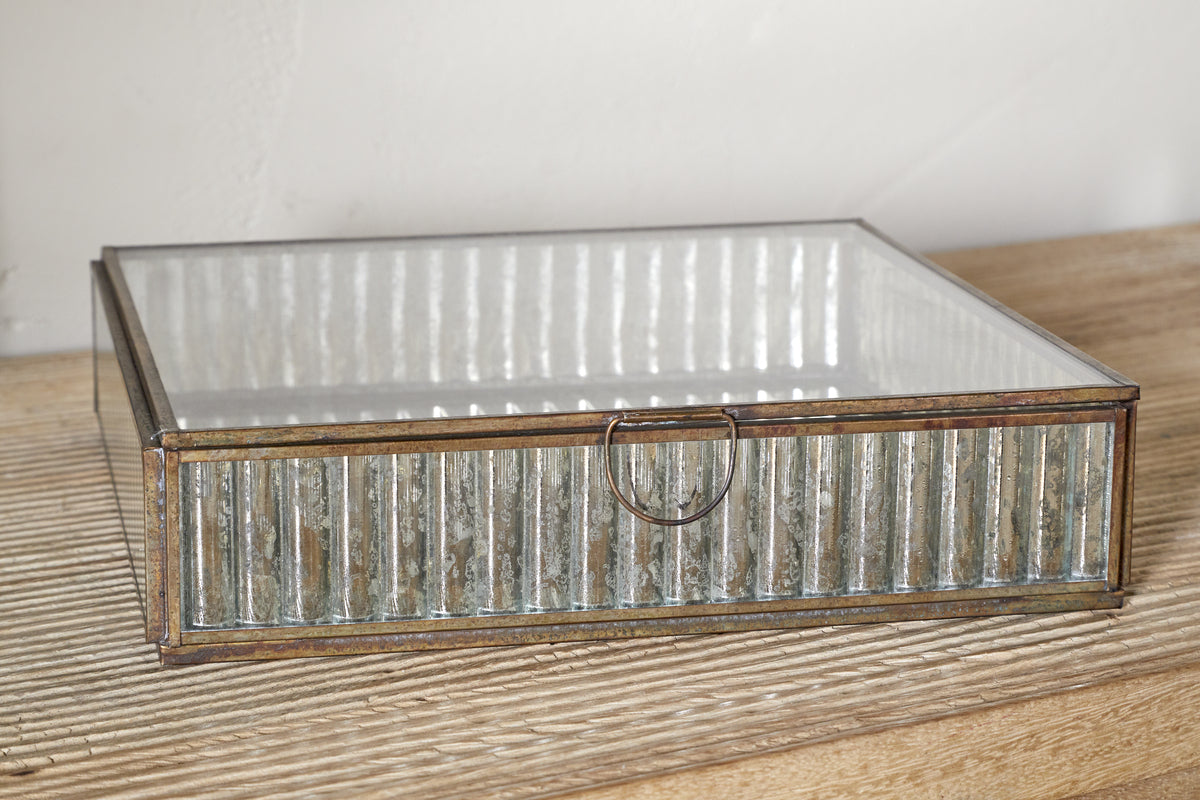 The nkuku Malita Jewellery Box features a gold-toned antique brass frame and ribbed mercury glass sides with a small metal ring pull, displayed on a light wood surface against a white wall.