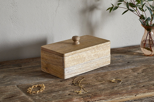 The nkuku Chanya Mango Wood Jewellery Box - Natural, featuring a round knob, sits on a rustic table. A gold bracelet and necklace are placed in front, with a glass vase of green foliage casting gentle shadows nearby.