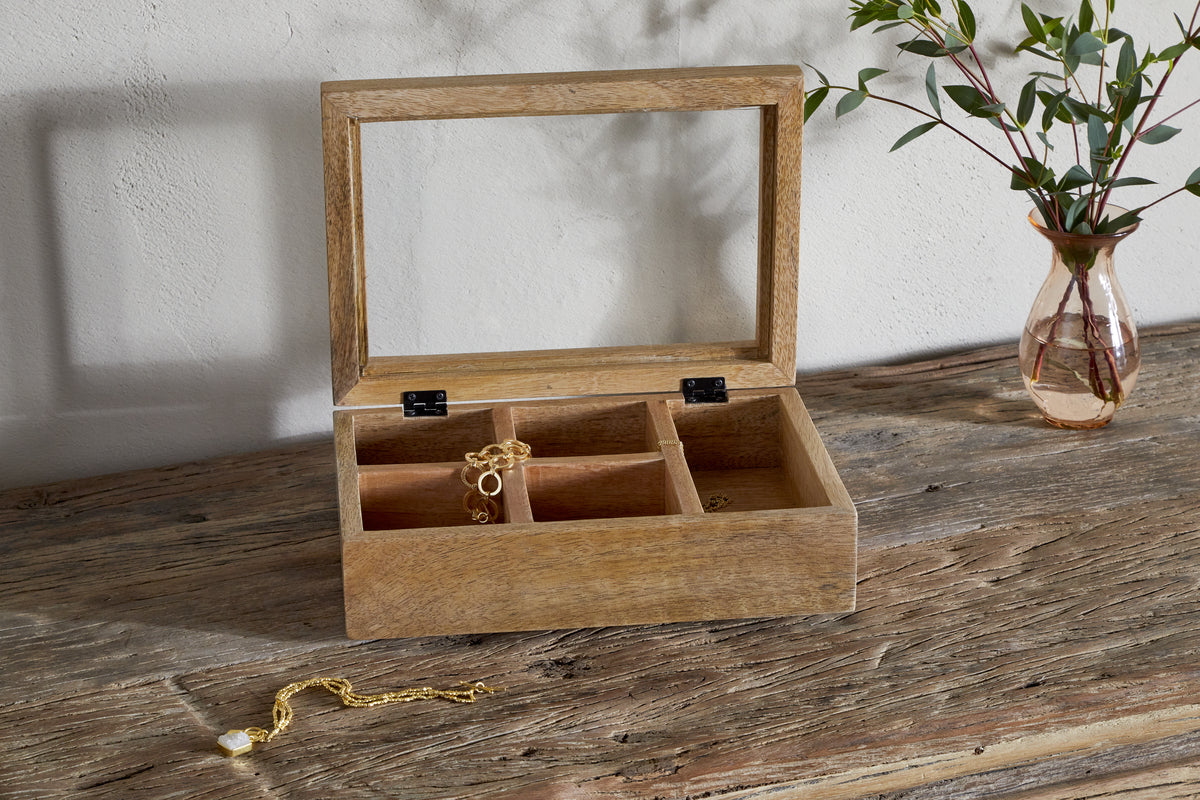 The nkuku Kabani Mango Wood Jewellery Box - Natural, featuring an open glass lid and four compartments with gold jewellery, sits on a rustic table. A gold bracelet with a pearl charm is outside, and a glass vase with leafy branches is behind.