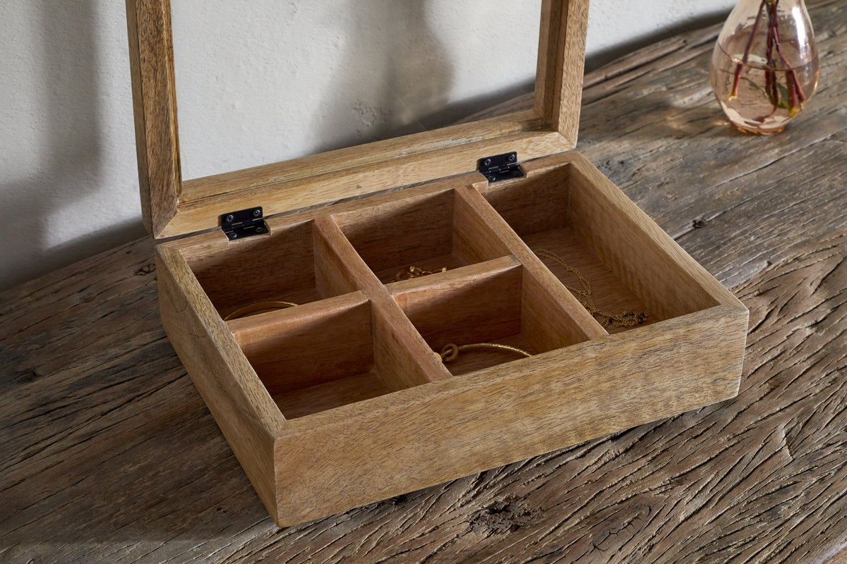 The nkuku Kabani Mango Wood Jewellery Box - Natural, with five compartments and an unfinished wood look, sits partially open on a rustic table near a clear vase holding fresh stems in water.