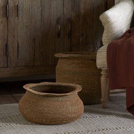 The nkuku Rana Short Baskets in Natural & Moss sit on a textured beige rug next to a wooden cabinet, while a light chair with a chunky rust-red knit blanket adds to the cozy, rustic look—ideal for fans of natural home decor.