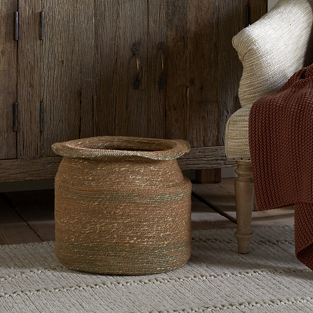 The nkuku Rana Tall Basket in Natural & Moss sits on a textured rug by a rustic wooden cabinet. Nearby, a wooden chair with a cream cushion and maroon-brown knit blanket adds to the cozy, earthy vibe.