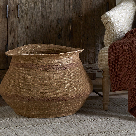 The nkuku Rana Wide Basket in Natural & Rust sits on a textured rug by a wooden cabinet, while a beige chair nearby is draped with a rust knit blanket, creating a cozy and rustic atmosphere.