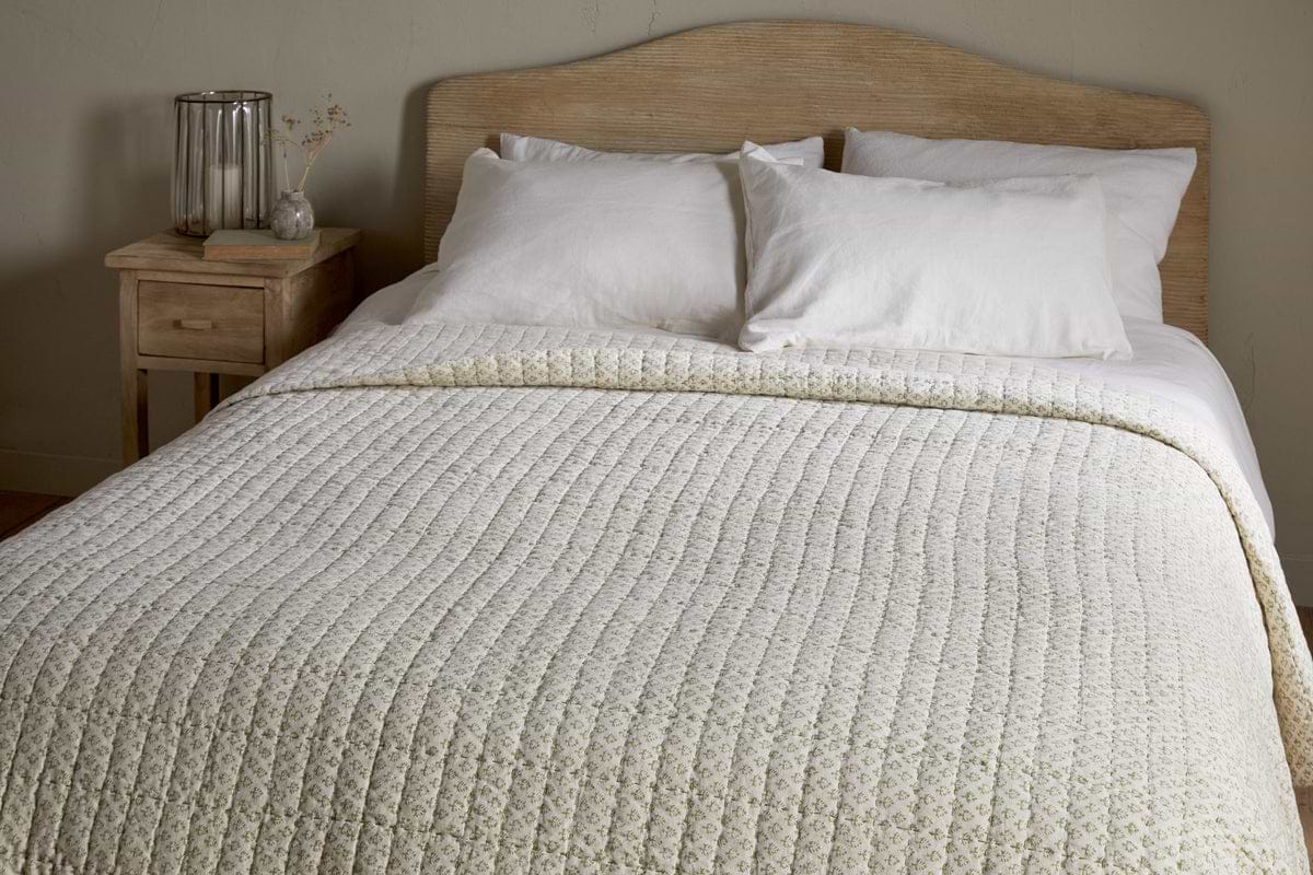 A neatly made bed with a light wood headboard features nkuku’s Peplu Cotton & Linen Screen Print Bed Quilt in Sage Green, two white pillows, and a small wooden nightstand holding a glass vase of dried flowers and a clear lamp in a neutral-toned room.