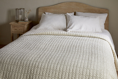 A neatly made bed with a light wood headboard features nkuku’s Peplu Cotton & Linen Screen Print Bed Quilt in Sage Green, two white pillows, and a small wooden nightstand holding a glass vase of dried flowers and a clear lamp in a neutral-toned room.