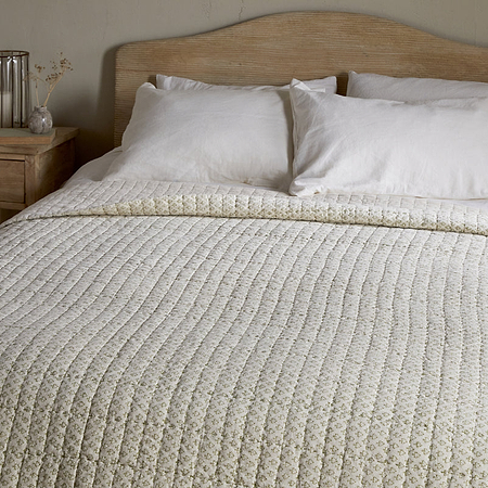A neatly made bed with a light wood headboard features nkuku’s Peplu Cotton & Linen Screen Print Bed Quilt in Sage Green, two white pillows, and a small wooden nightstand holding a glass vase of dried flowers and a clear lamp in a neutral-toned room.