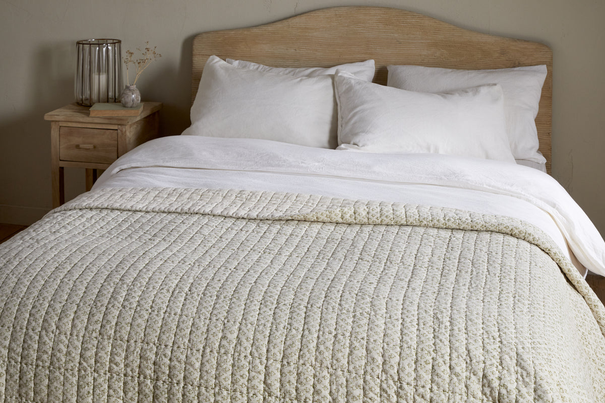 A neatly made bed with white pillows and sheets features the nkuku Peplu Cotton & Linen Screen Print Bed Quilt in sage green. A light wooden headboard and a nightstand with a glass vase, dried flowers, and decor stand against a neutral wall.