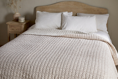 A neatly made bed features the nkuku Peplu Cotton & Linen Screen Print Bed Quilt in rust, three white pillows, and a wooden headboard. A wooden nightstand with a vase of dried flowers and decor accents stands by a neutral wall.