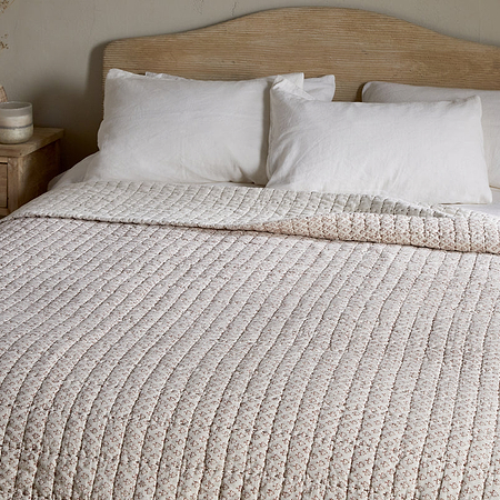 A neatly made bed features the nkuku Peplu Cotton & Linen Screen Print Bed Quilt in rust, three white pillows, and a wooden headboard. A wooden nightstand with a vase of dried flowers and decor accents stands by a neutral wall.