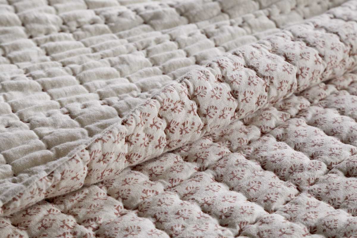 A close-up of the nkuku Peplu Cotton & Linen Screen Print Bed Quilt in Rust shows a folded corner, highlighting its soft, textured surface and intricate repeating pattern—showcasing handmade Indian artistry and cozy appeal.