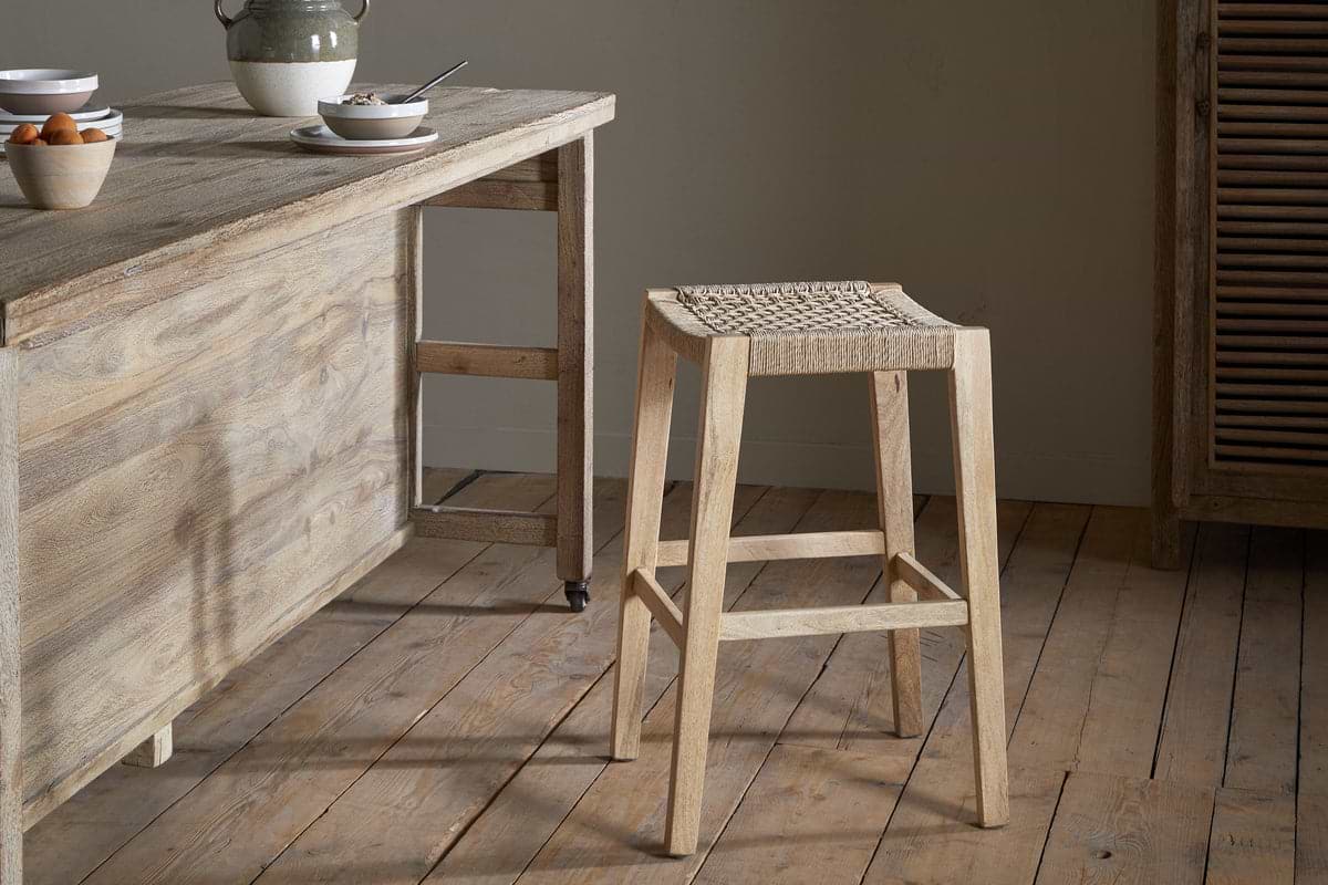 A natural wood kitchen with a rustic breakfast table, set with bowls and a jug, is complemented by the nkuku Aamani Mango Wood & Cord Counter Stool - Natural, featuring a woven seat. Wooden floors and soft neutral lighting complete the scene.