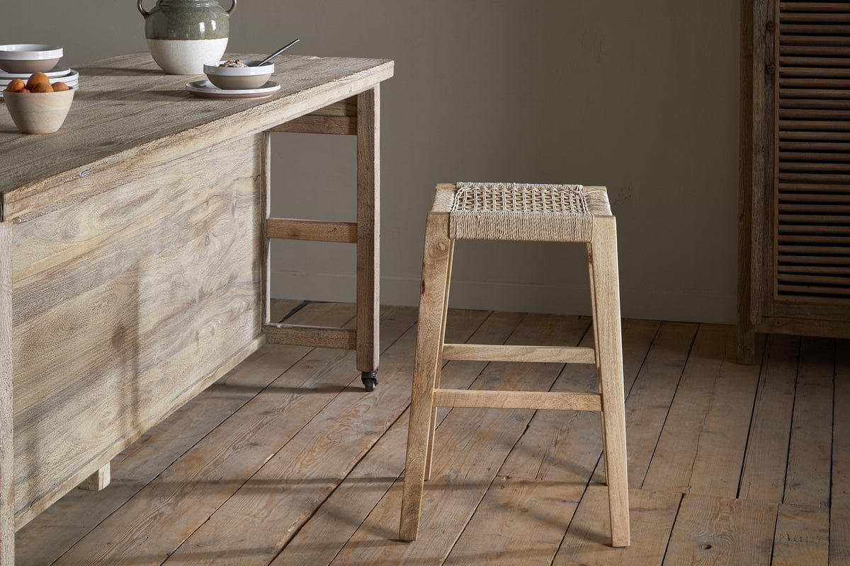 A rustic kitchen shows a light mango wood table set for breakfast and the Aamani Mango Wood & Cord Counter Stool by nkuku, featuring a woven seat in natural tones. The wide plank floor and neutral backdrop complete the scene.
