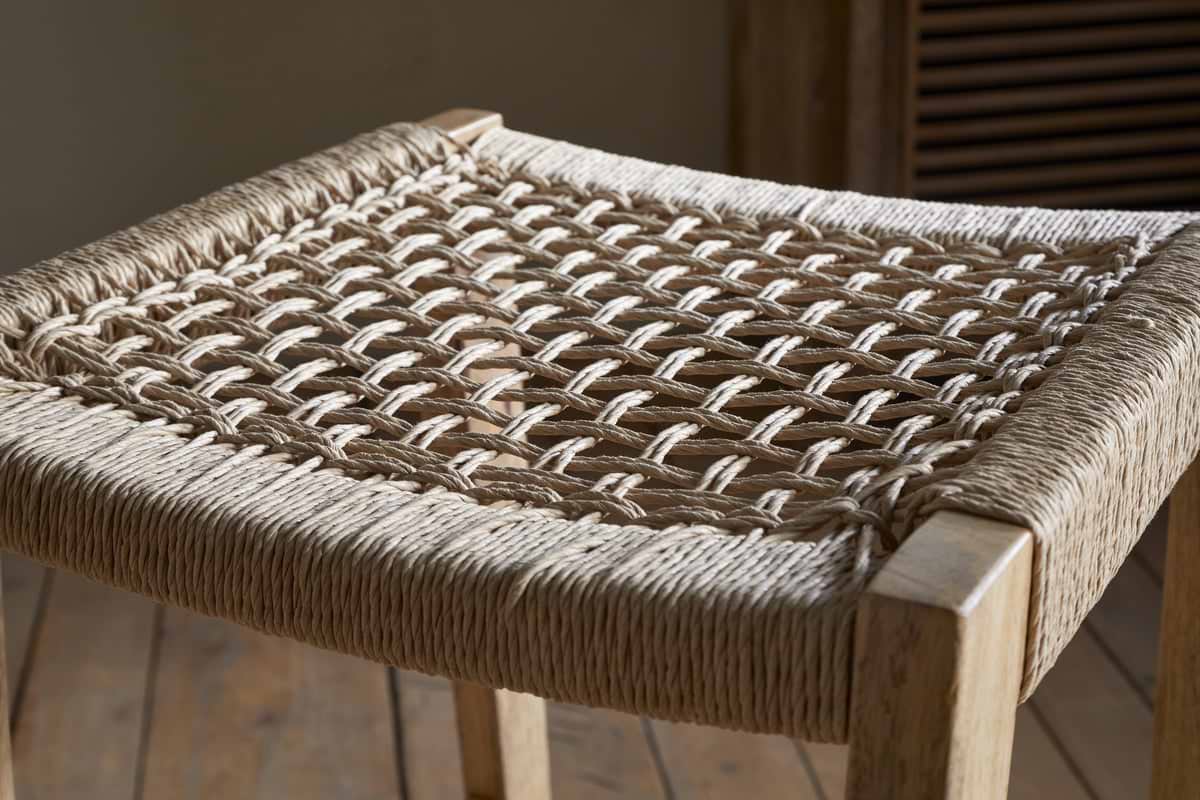A close-up of the nkuku Aamani Mango Wood & Cord Counter Stool - Natural reveals its sturdy mango wood frame and open, crisscross woven seat crafted from thick, natural cord, set against a wooden floor with a heating vent in the background.