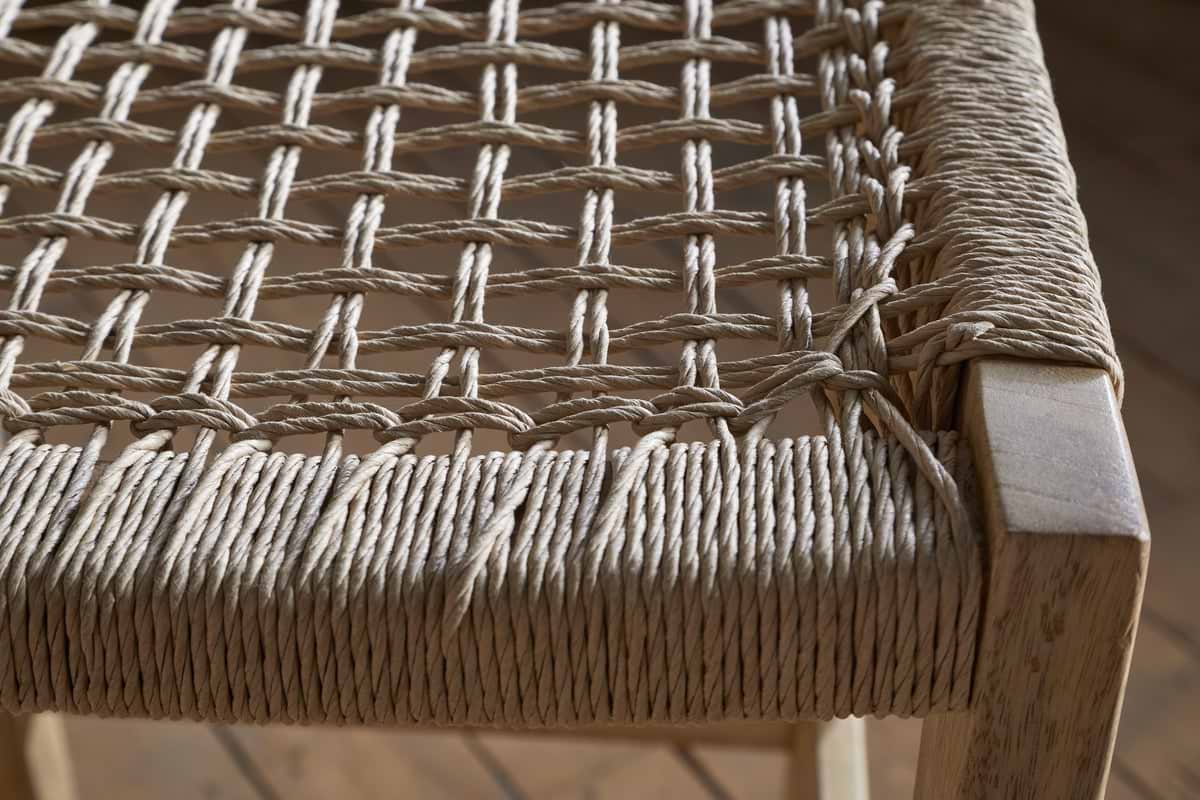Close-up of the nkuku Aamani Mango Wood & Cord Counter Stool - Natural, featuring a thick, natural-colored rope seat woven in a crisscross pattern on a light mango wood frame. The detailed craftsmanship and texture are clearly visible. Wooden floor background.