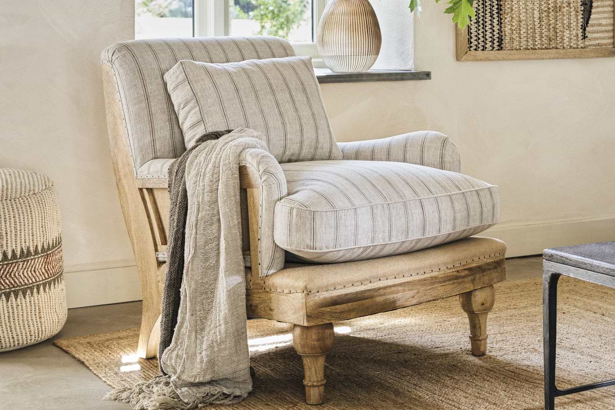 The nkuku Abe Deconstructed Linen Armchair in Natural Stripe features a mango wood frame and comes with a matching pillow and throw, creating a cozy spot on a woven rug. A window, decorative vase, and textured wall art add warmth to the inviting room.