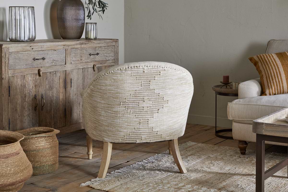 A warm, inviting living room with the nkuku Aranya Cotton & Jute Occasional Chair in natural beige, a rustic wooden sideboard with ribbed glass vases, woven baskets on wood flooring, a cream sofa with striped cushions, and a textured cream rug.