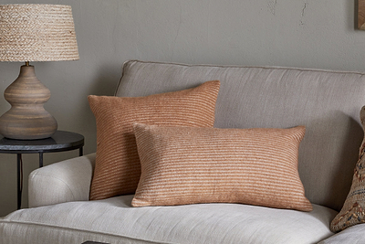 A beige sofa is paired with two Ilaa Wool Cushion Covers in Sand by nkuku, featuring rust-colored stripes. Beside it stands a round dark side table with a textured lamp, set against a neutral light gray wall for a cozy, minimalist look.