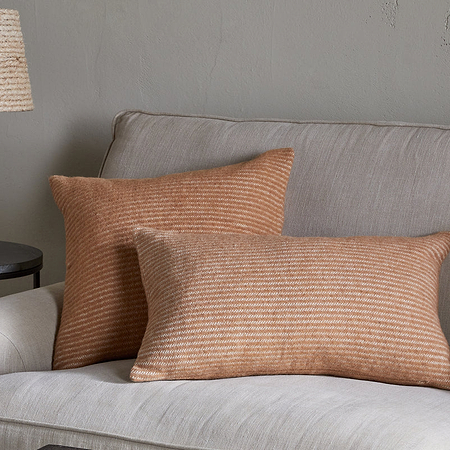 A beige sofa is paired with two Ilaa Wool Cushion Covers in Sand by nkuku, featuring rust-colored stripes. Beside it stands a round dark side table with a textured lamp, set against a neutral light gray wall for a cozy, minimalist look.