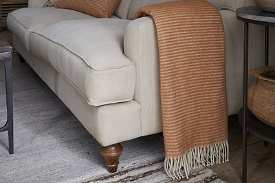 A beige upholstered sofa from the Ilaa collection with wooden legs is draped with the nkuku Ilaa Wool Throw in Sand, featuring textured tan-and-white stripes. The sofa sits on a soft gray rug, with a woven basket and round table nearby.