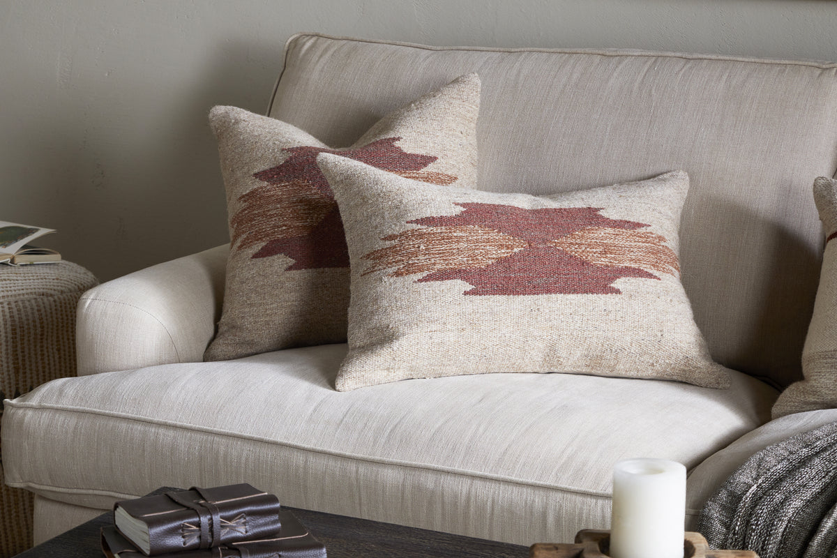 Two beige throw pillows, one in the nkuku Kabir Wool Cushion Cover - Rust, featuring a southwestern-style geometric design, rest on a light beige sofa. A textured tan blanket drapes the armrest; a coffee table holds a candle and book.