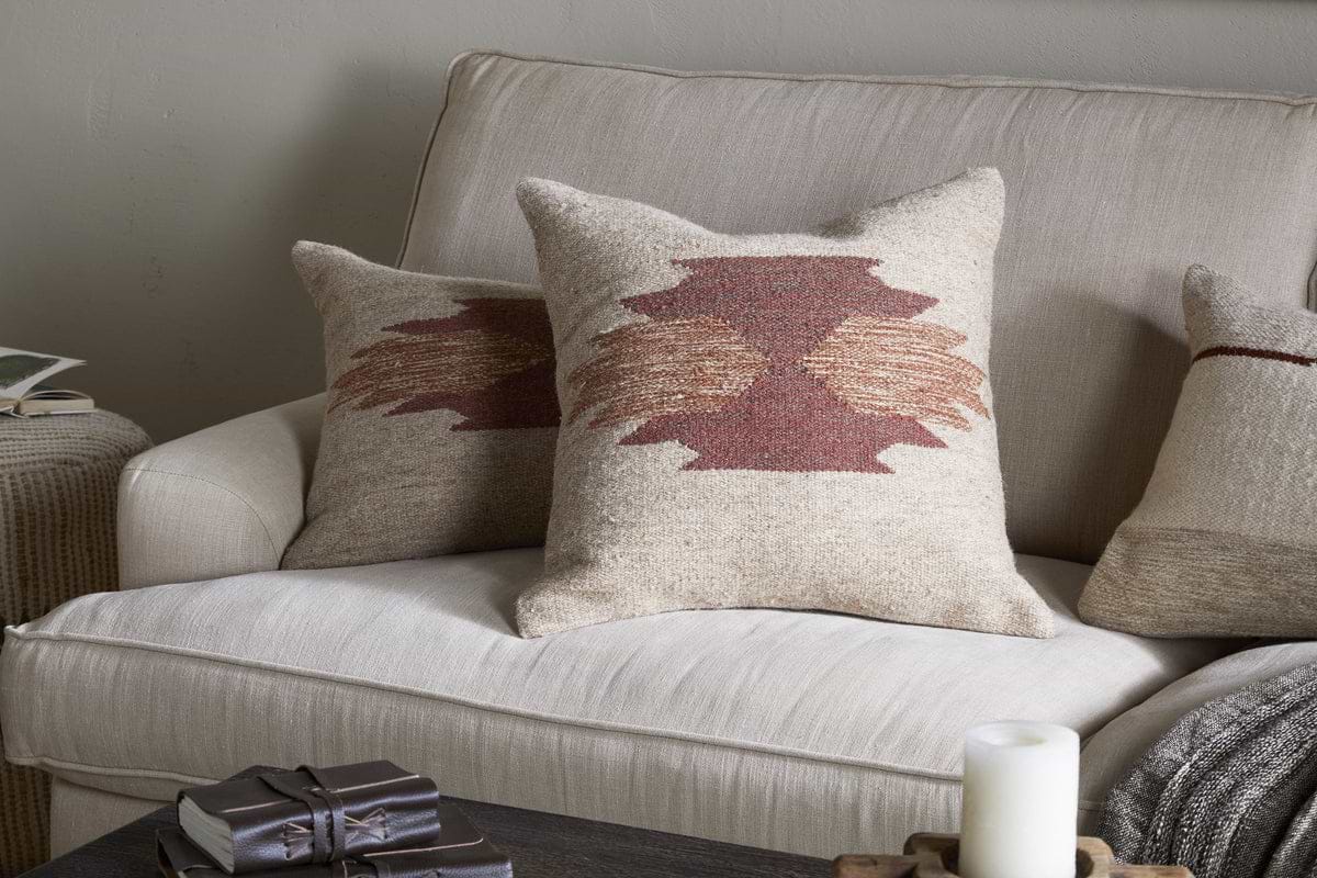 Two Kabir Wool Cushion Covers - Rust by nkuku, featuring geometric brown and rust patterns, rest on a tan sofa. Paired with a brown blanket, decorative cushions, a dark wooden coffee table, candle, and books for a cozy neutral look.