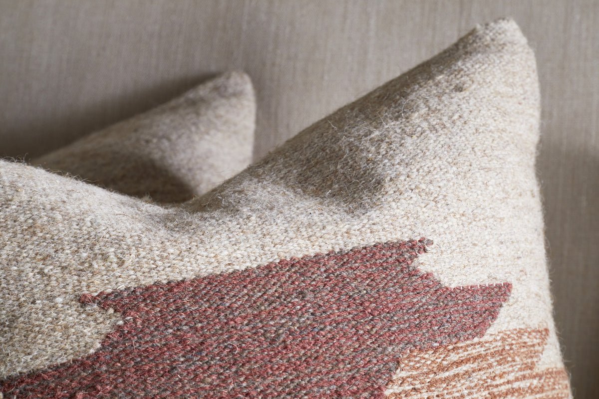 The Kabir Wool Cushion Cover - Rust from nkuku features a textured, geometric southwestern-inspired design in earthy tones. Made of a cotton and wool blend, it sits against a neutral background with another similar pillow behind it.