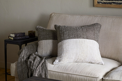 A beige sofa with two square pillows, one in the nkuku Munir Linen Cushion Cover—charcoal with gray and off-white color blocks. A gray knit throw rests on the armrest; nearby, a dark side table holds stacked books and a mug against a light wall.