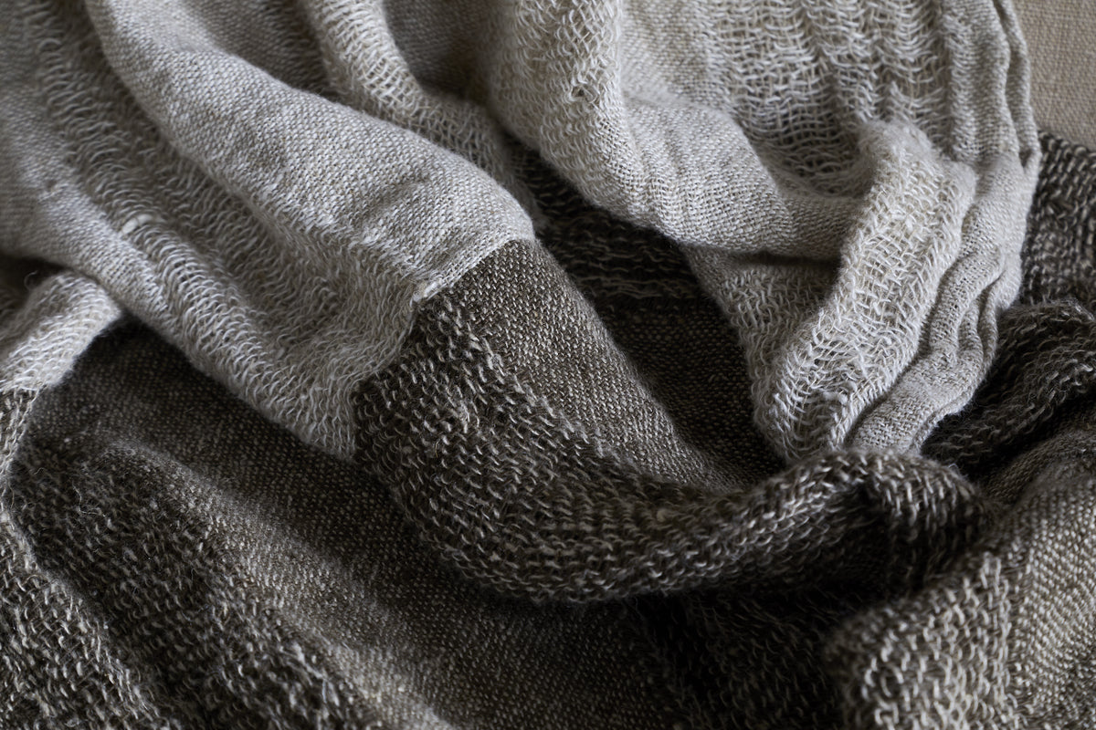 A close-up of the Munir Linen Throw in Charcoal by nkuku shows soft, textured wavy folds and wide alternating stripes in light beige and dark brown, highlighting its intricate knit pattern and cozy appeal.