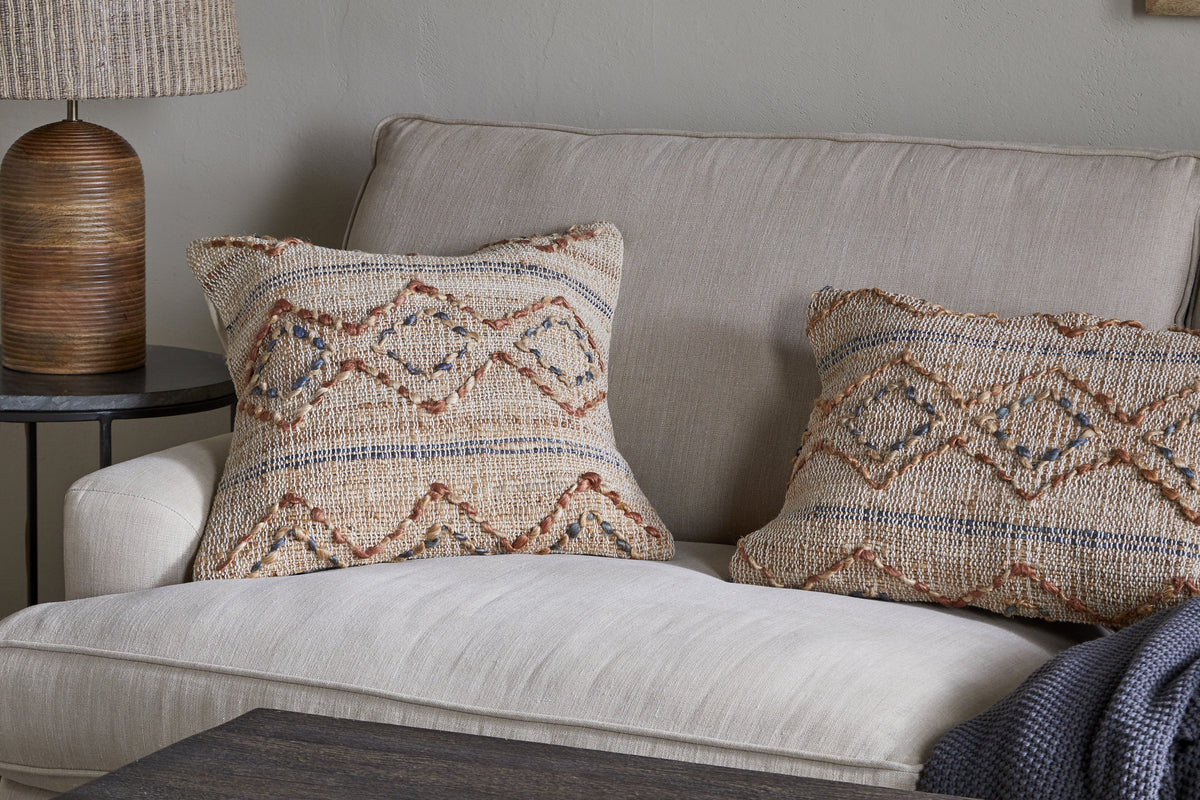 A beige sofa with two patterned throw pillows—one featuring the nkuku Nila Esha Cushion Cover in Rust & Indigo—sits by a round black side table topped with a ribbed brown lamp. A dark blue knit blanket is draped over the arm against a light gray wall.