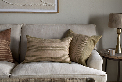A beige sofa with an nkuku Eesha Jute Cushion Cover in sage green and two earth-toned striped pillows sits by a light wall. A framed artwork hangs above, while a round side table with a beige lamp and cup adds to the modern rustic style.
