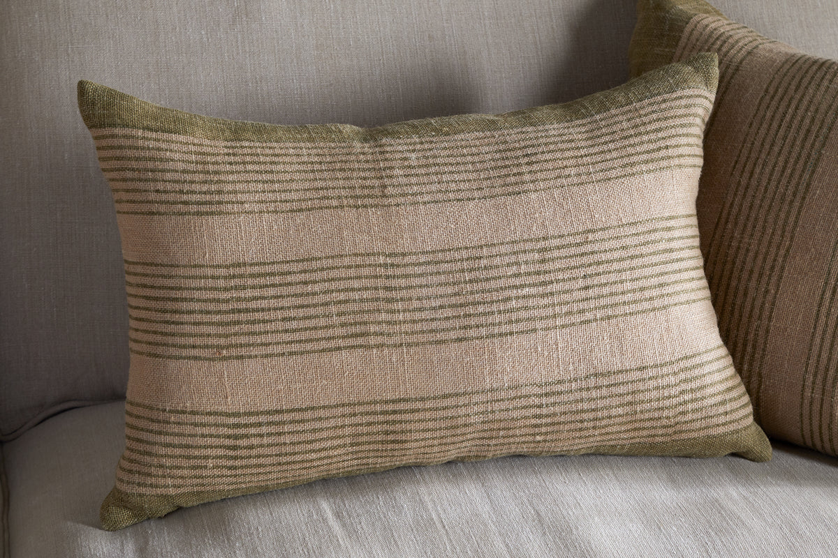 The Eesha Jute Cushion Cover - Sage Green by nkuku rests on a light gray fabric sofa, bringing modern rustic charm. Its soft, woven texture and striped design pair perfectly with a similar block-printed cushion cover behind it.