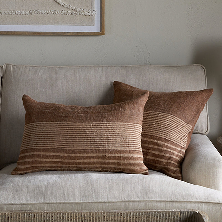 A cozy beige sofa is styled with modern rustic touches, including nkuku’s Eesha Jute Cushion Cover in Rust, brown striped and green pillows, a round wooden table with a lamp and candle, and textured wall art above in neutral tones.