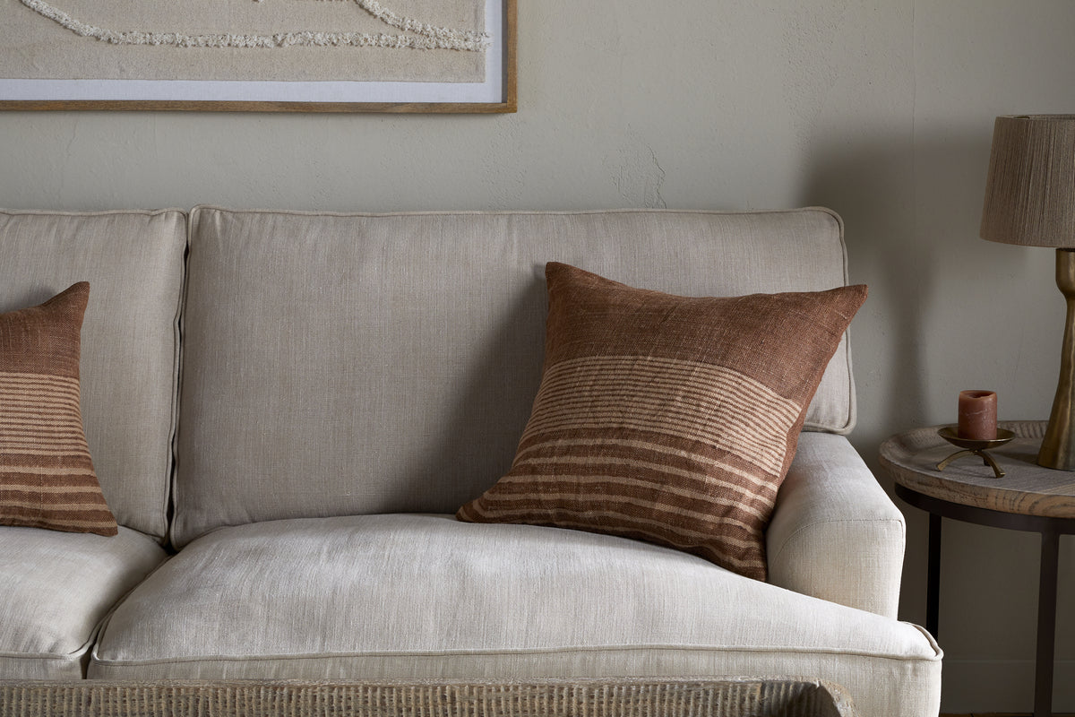 A beige fabric sofa is styled with two nkuku Eesha Jute Cushion Covers in Rust, featuring brown and tan stripes. Against a light wall, the cozy scene is completed with framed art and a round wooden side table topped with a lamp and candle.