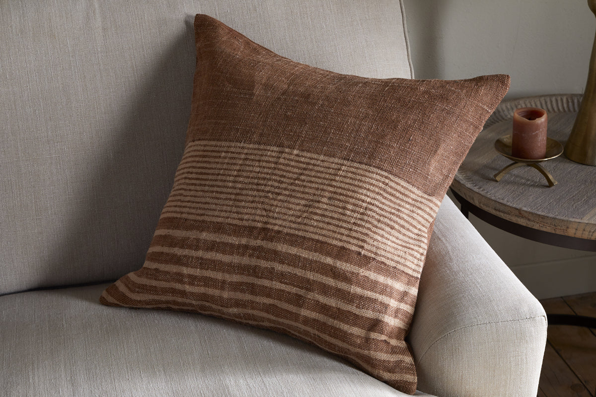 The Eesha Jute Cushion Cover in rust by nkuku, featuring modern rustic woven stripes, sits on a light gray sofa. A round wooden table with a brass lamp and brown candle in the background adds to the cozy ambiance.