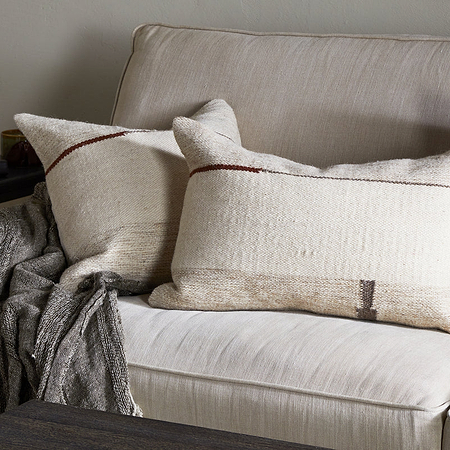 A cozy beige sofa features two nkuku Tamil Wool Cushion Covers in natural, with simple brown, charcoal, and rust tones. A gray woven throw is draped over the arm, and a side table with stacked books completes this warm, inviting setting.