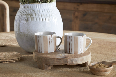 Two small Patthar Mugs in Cream & Clay by nkuku sit on a rustic wooden stand atop a table. Behind them, a white textured vase with greenery and woven coasters add charm to this warmly lit, dinnerware-inspired scene.