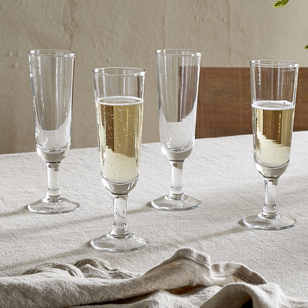 Four Pani Champagne Flutes by nkuku, in a set of 4 clear glasses, are arranged on a beige tablecloth; two are filled with bubbly champagne and two are empty. A folded napkin rests nearby alongside a striped bowl and leafy branch.