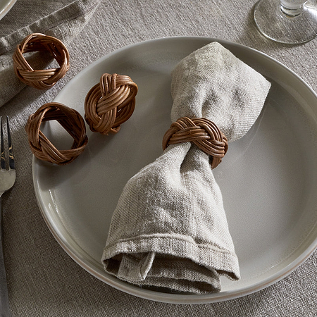 On a white plate sits a beige linen napkin with an nkuku Bersama Rattan Napkin Ring in Natural; two extra rings from the set of 4 rest nearby. Utensils and a glass complete the elegant look atop a neutral linen tablecloth.