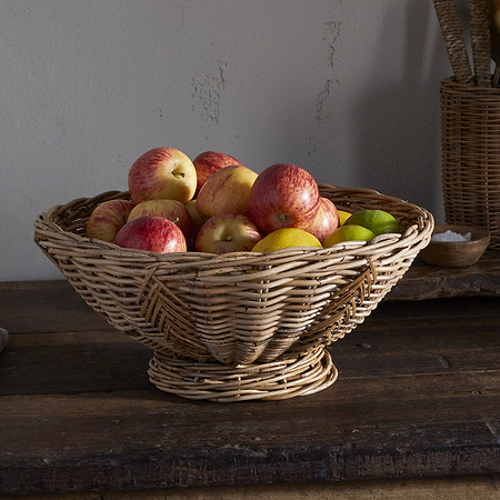 The Bersama Rattan Fruit Bowl by nkuku, filled with apples and a lime, sits on a rustic wooden table—a charming kitchen centerpiece with utensils in a wicker holder and a folded cloth in the background.