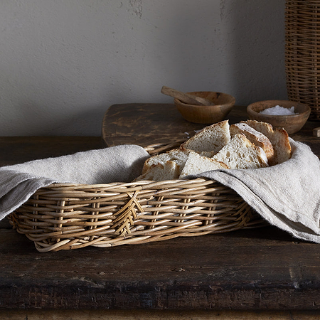 The nkuku Bersama Rattan Bread Basket - Natural, lined with beige cloth, displays rustic bread on a wooden table. Stacked white bowls and wooden salt bowls complete the cozy, rustic kitchen decor in the background.
